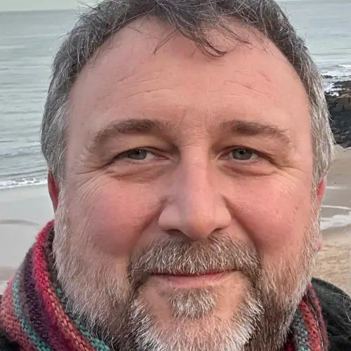 Craig Humble, Owner of Art of Protest Gallery smiling with grey hair and beard wearing a multicolored scarf near a beach.