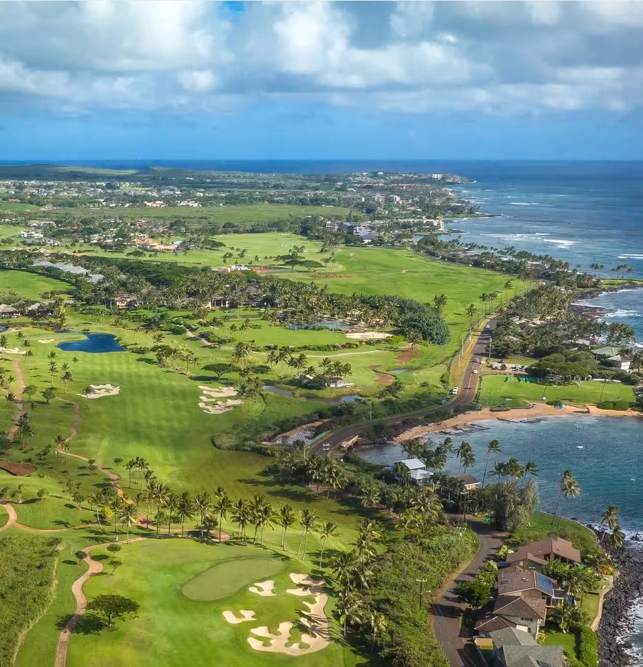 Aerial view of a coastal golf course with green fairways, palm trees, a road, beachfront houses, and the ocean under a partly cloudy sky.