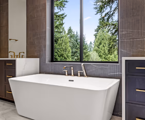 Modern bathroom with a white freestanding bathtub beneath a large window showing green trees outside.