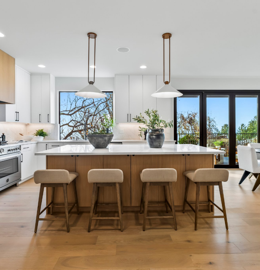 Modern kitchen with a large white island, four wooden stools, pendant lights, white cabinets, and large windows showing outdoor trees.