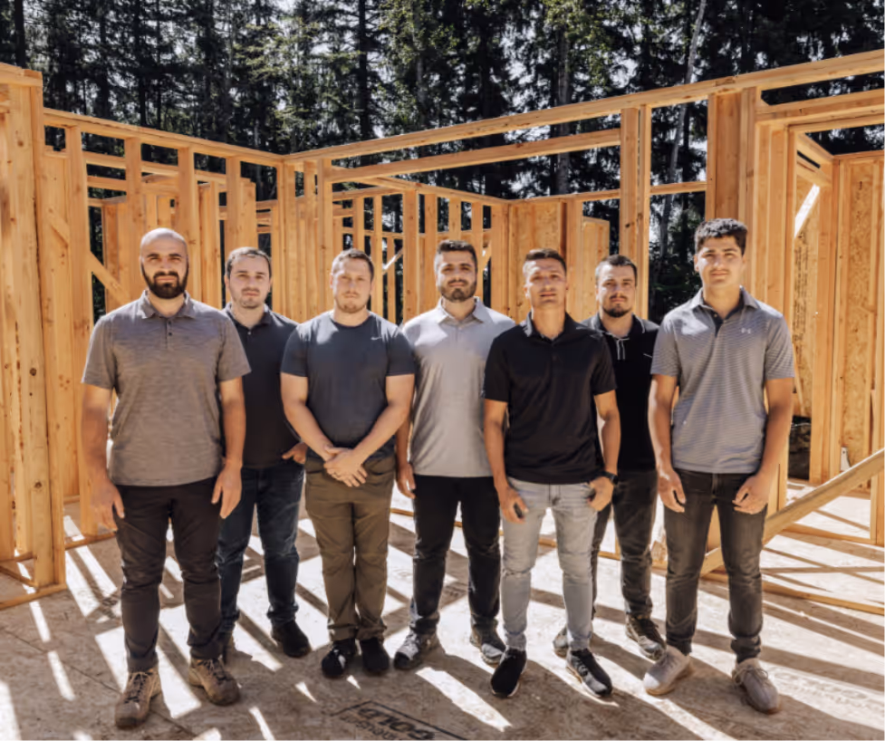Seven men standing in a row inside a wooden frame structure of a building under construction with trees in the background.
