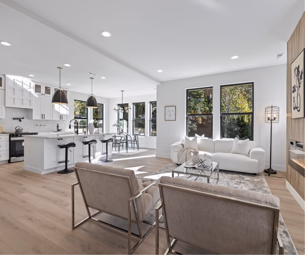 Bright open-concept living room and kitchen with white furniture, large windows, wooden floor, and modern lighting.