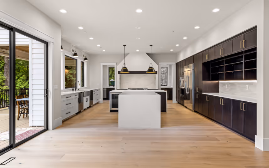 Spacious modern kitchen with light wood flooring, white countertops, dark wood cabinetry, two pendant lights over a central island, and large sliding glass doors to an outdoor patio.