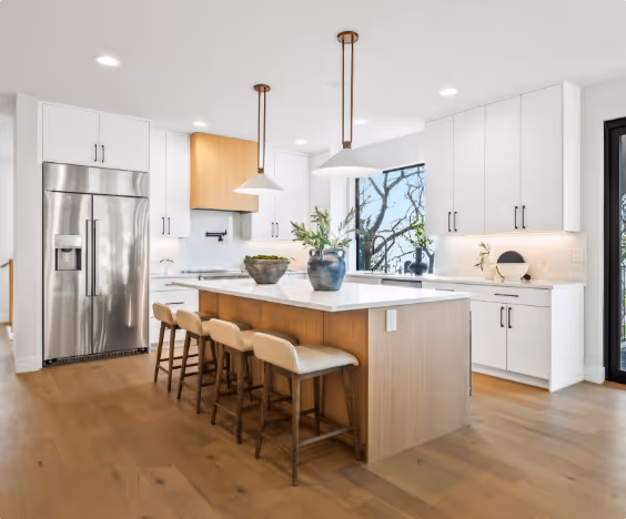 Modern kitchen with wooden floor, large island with four chairs, white cabinets, stainless steel refrigerator, and two pendant lights.