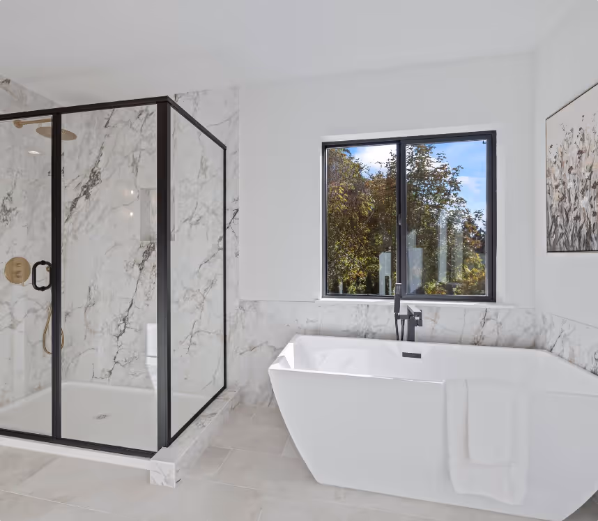Modern bathroom with a freestanding white bathtub, a black-framed glass shower enclosure, marble walls, and a window showing greenery outside.