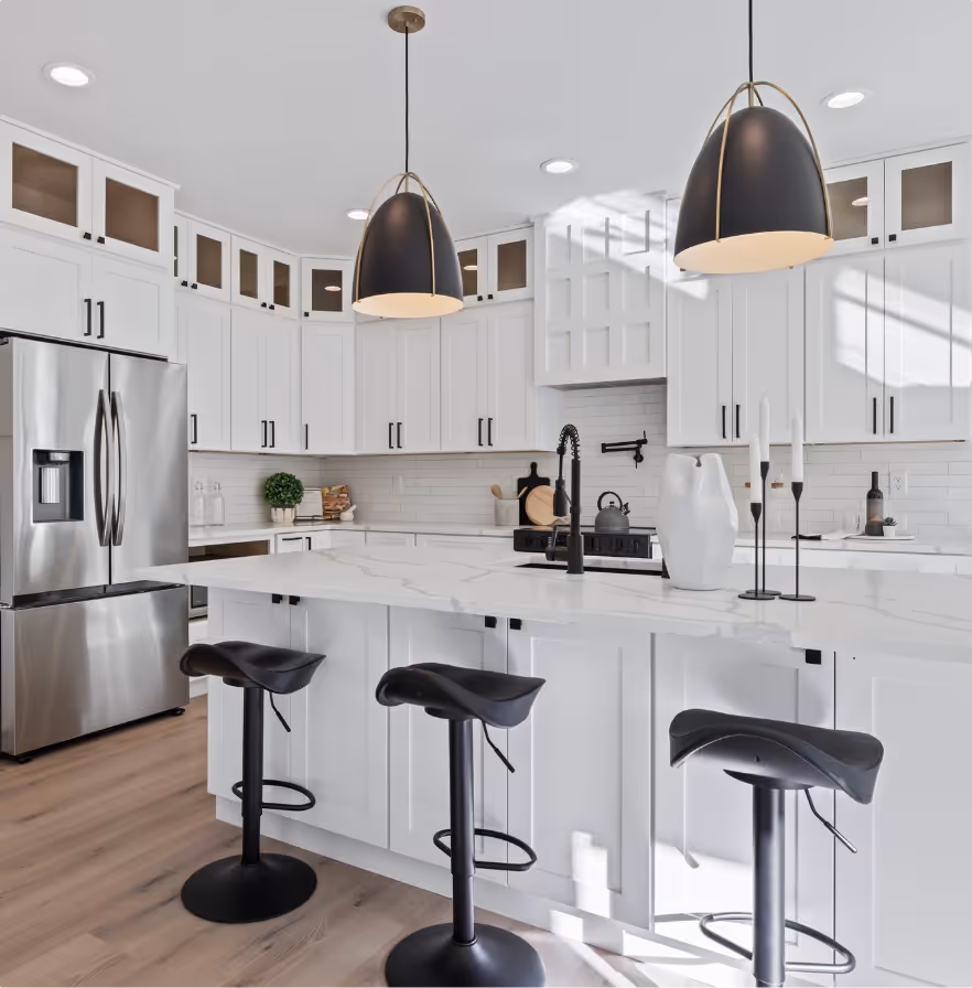 Modern kitchen with white cabinets, marble countertop island, black bar stools, two pendant lights, and stainless steel refrigerator.