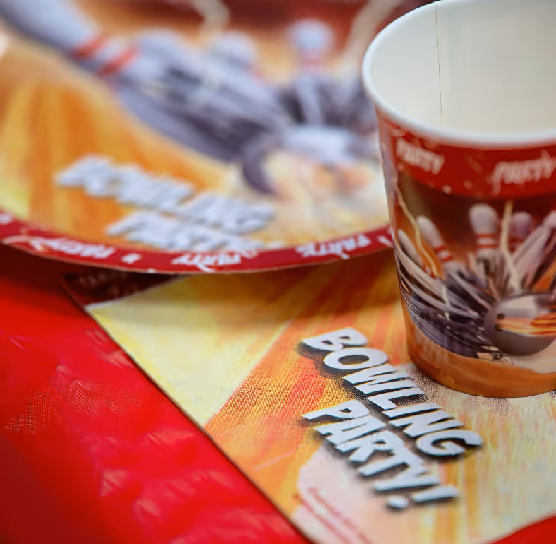 Paper cup and party plate with bowling pins and ball design placed on a surface with 'Bowling Party!' text.