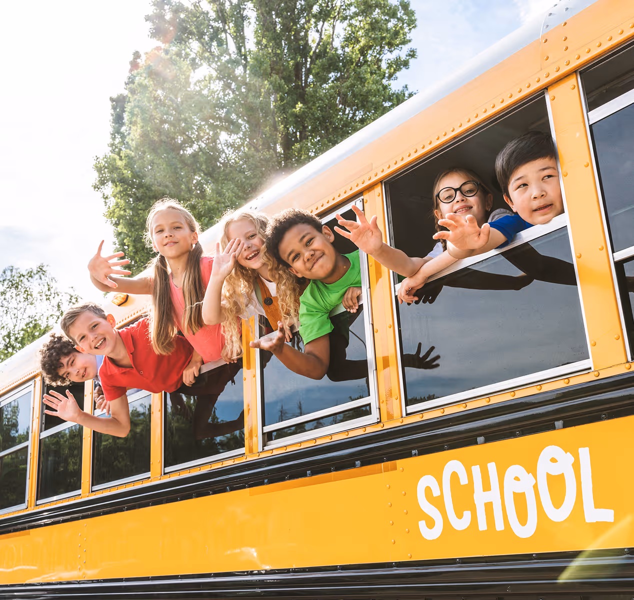 Six children smiling and waving from the windows of a yellow school bus on a sunny day.