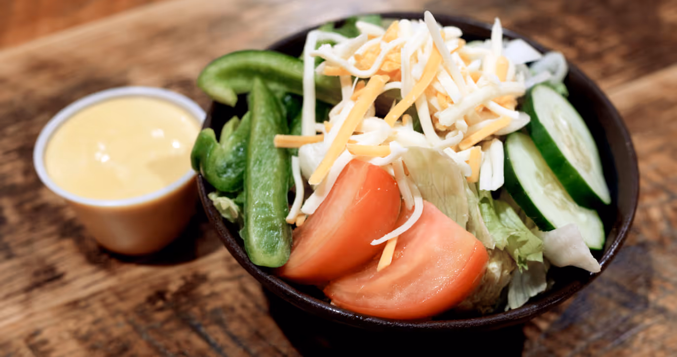 Bowl of tossed salad with lettuce, tomato wedges, sliced cucumber, green bell pepper, shredded cheese, and a side of creamy dressing.