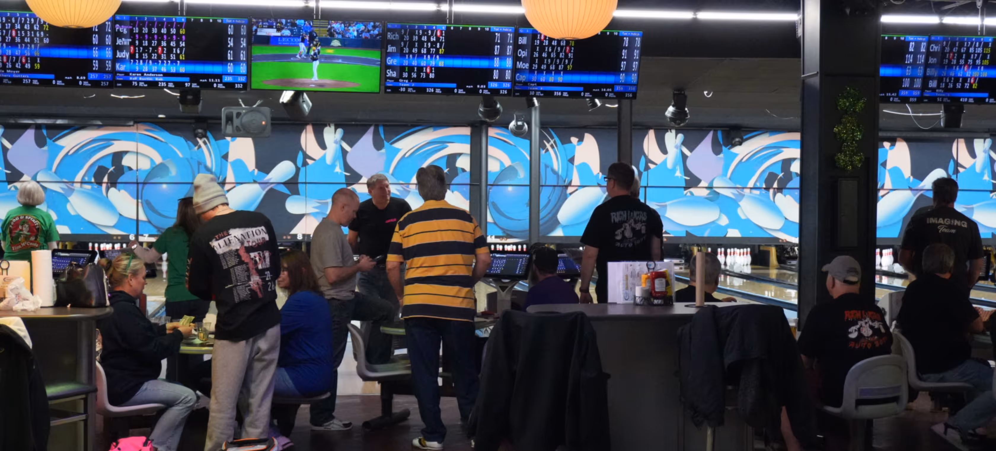 People socializing and bowling inside a lively bowling alley with animated screens and scoreboard displays above the lanes.