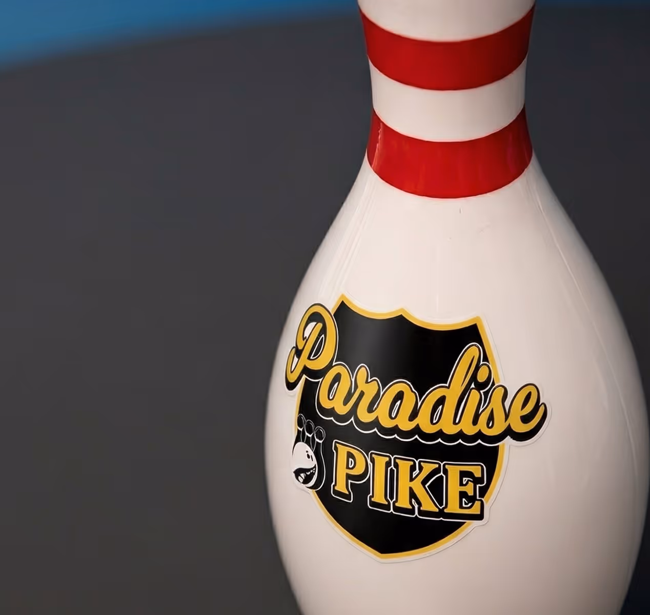 White bowling pin with red stripes and a black and gold 'Paradise Pike' logo.