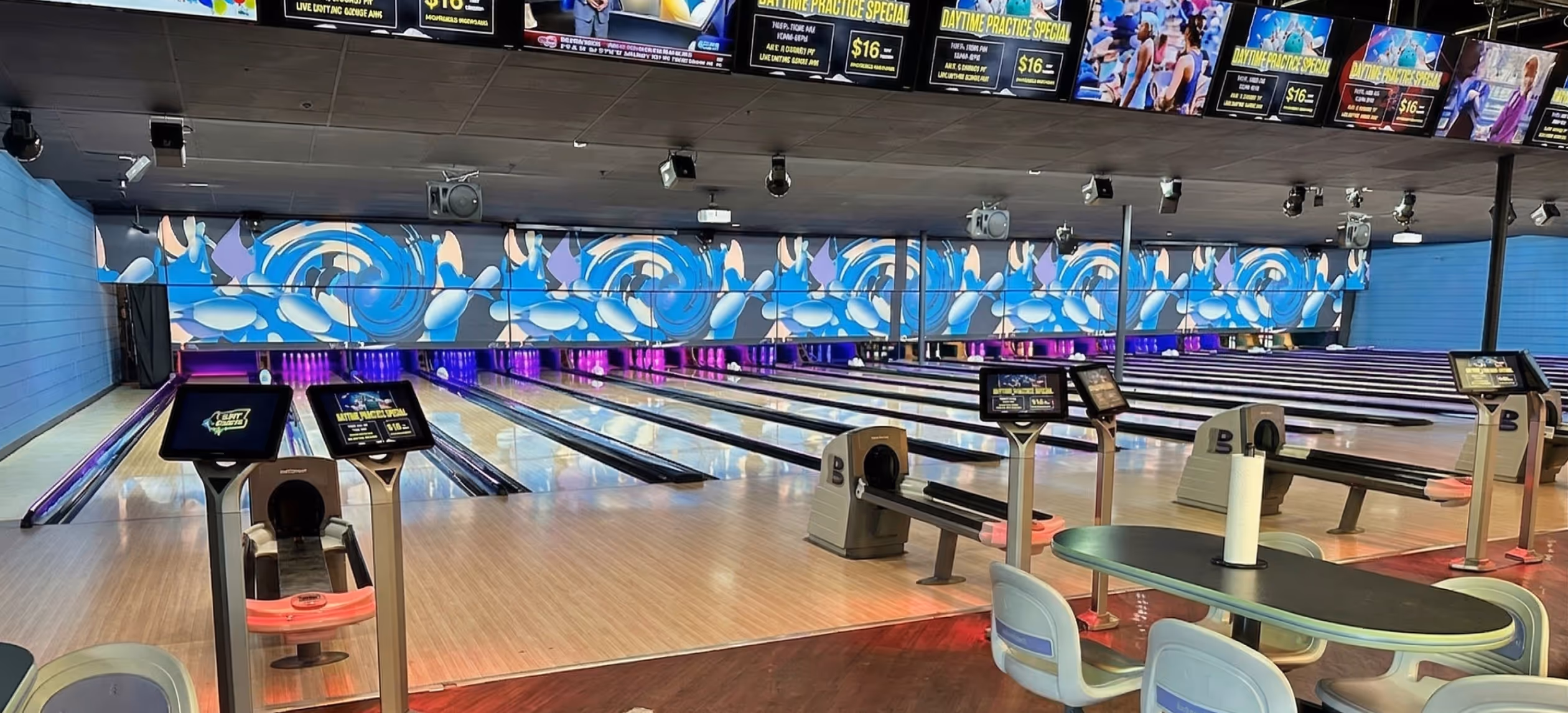 Indoor bowling alley with multiple lanes, digital scoring screens, light wood flooring, and seating area with a table and chairs.
