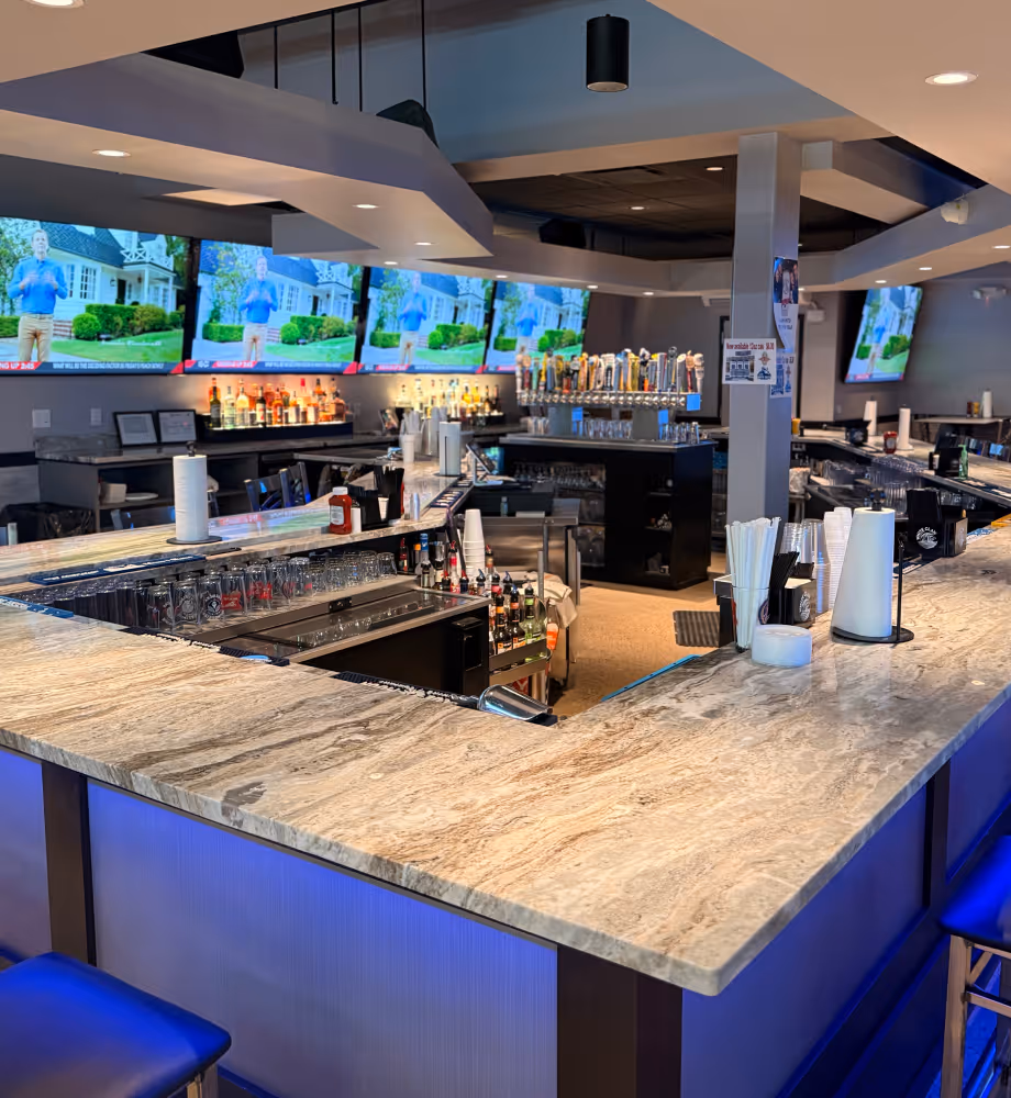 Modern bar interior with marble countertop, multiple TVs on the wall, and a variety of liquor bottles and beer taps visible.