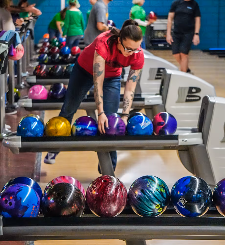 A woman with tattoos and glasses selecting a bowling ball from a rack in a bowling alley with other players in the background.