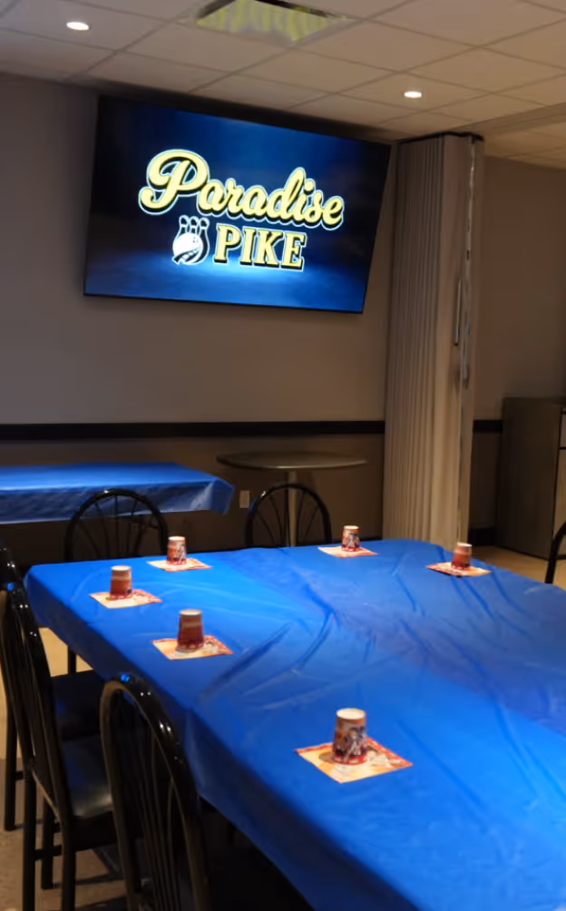 Room setup with tables covered in blue plastic tablecloths and small cups on napkins, with a screen on the wall displaying 'Paradise Pike' bowling logo.