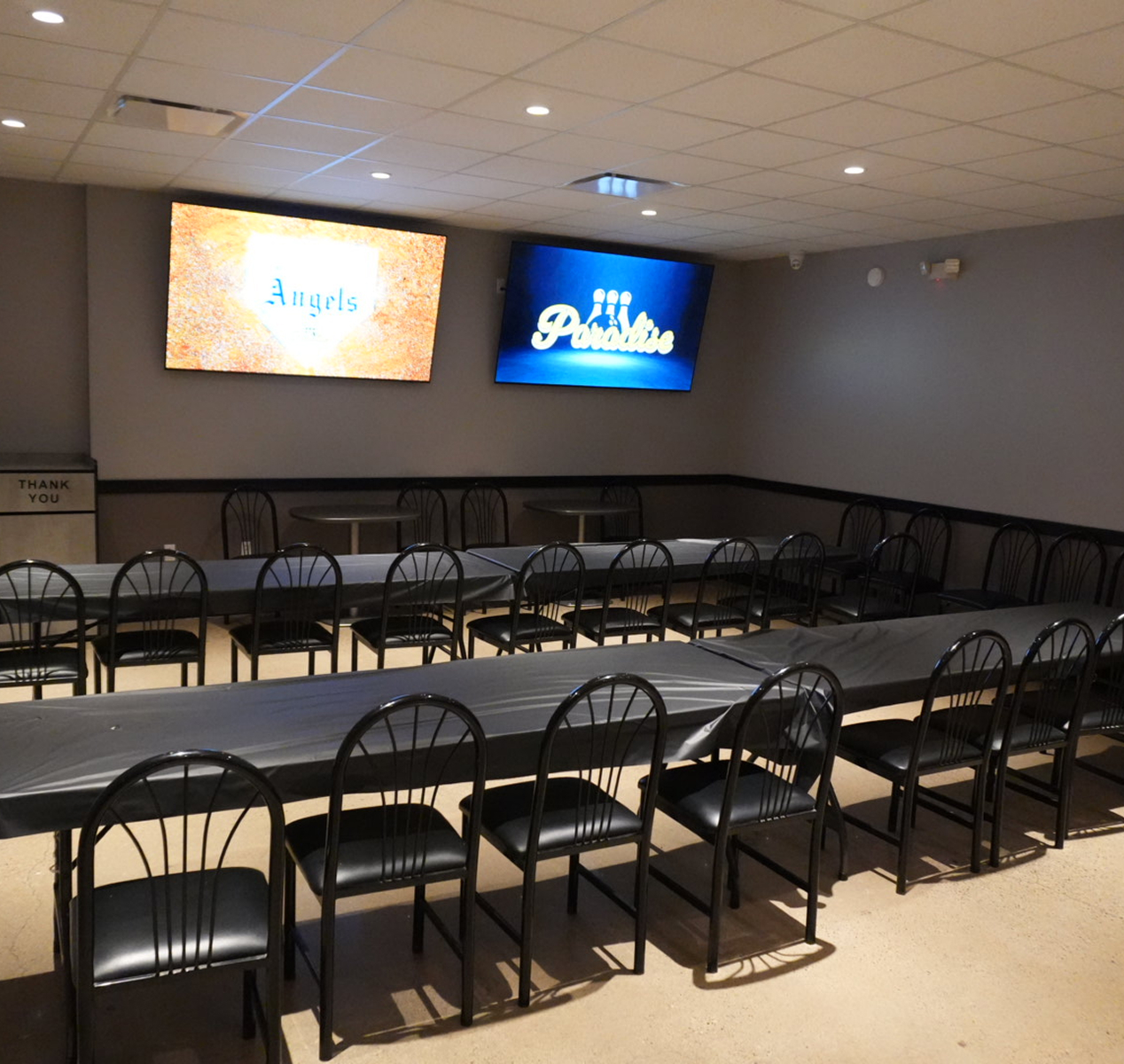 Empty event room with rows of black chairs and tables covered in black tablecloths, two wall-mounted TVs displaying 'Angels' and 'Paradise'.