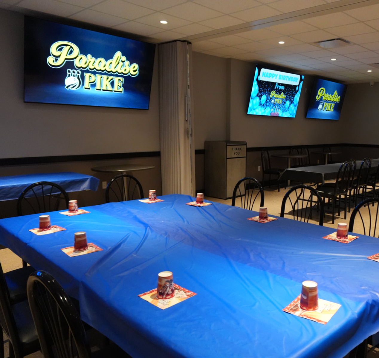 Party room with tables covered in blue plastic tablecloths and cups placed on napkins, three screens on the wall displaying 'Paradise Pike' and a birthday greeting.