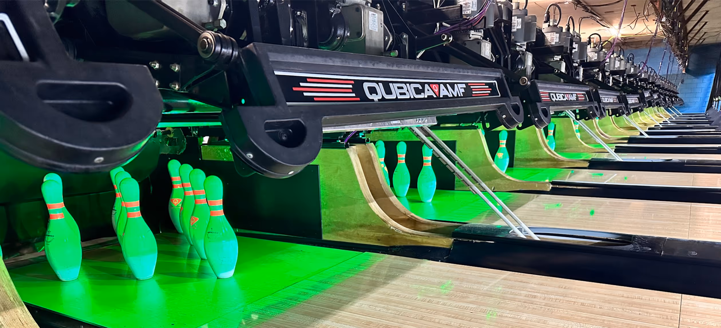 A row of bowling lanes with QubicaAMF pinsetters and green-lit bowling pins set up for a game.
