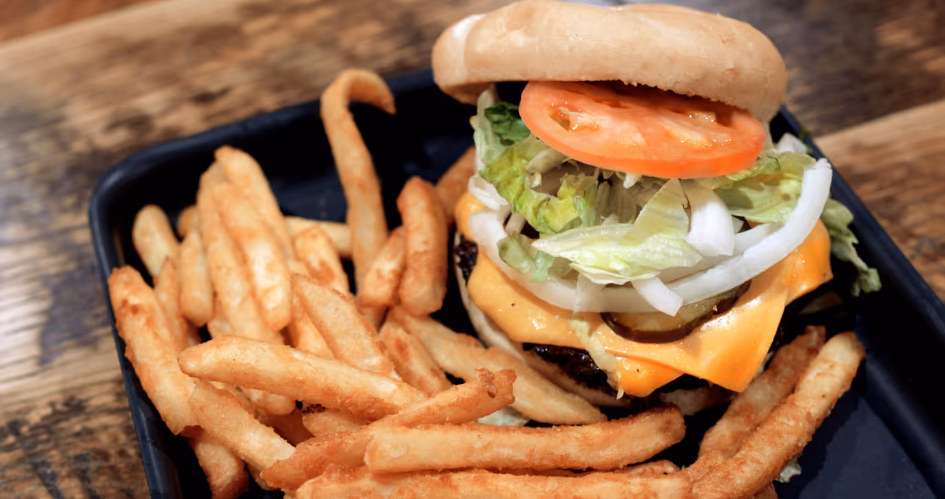 Cheeseburger with lettuce, tomato, onions, pickles, and melted cheese served with seasoned French fries on a black tray.