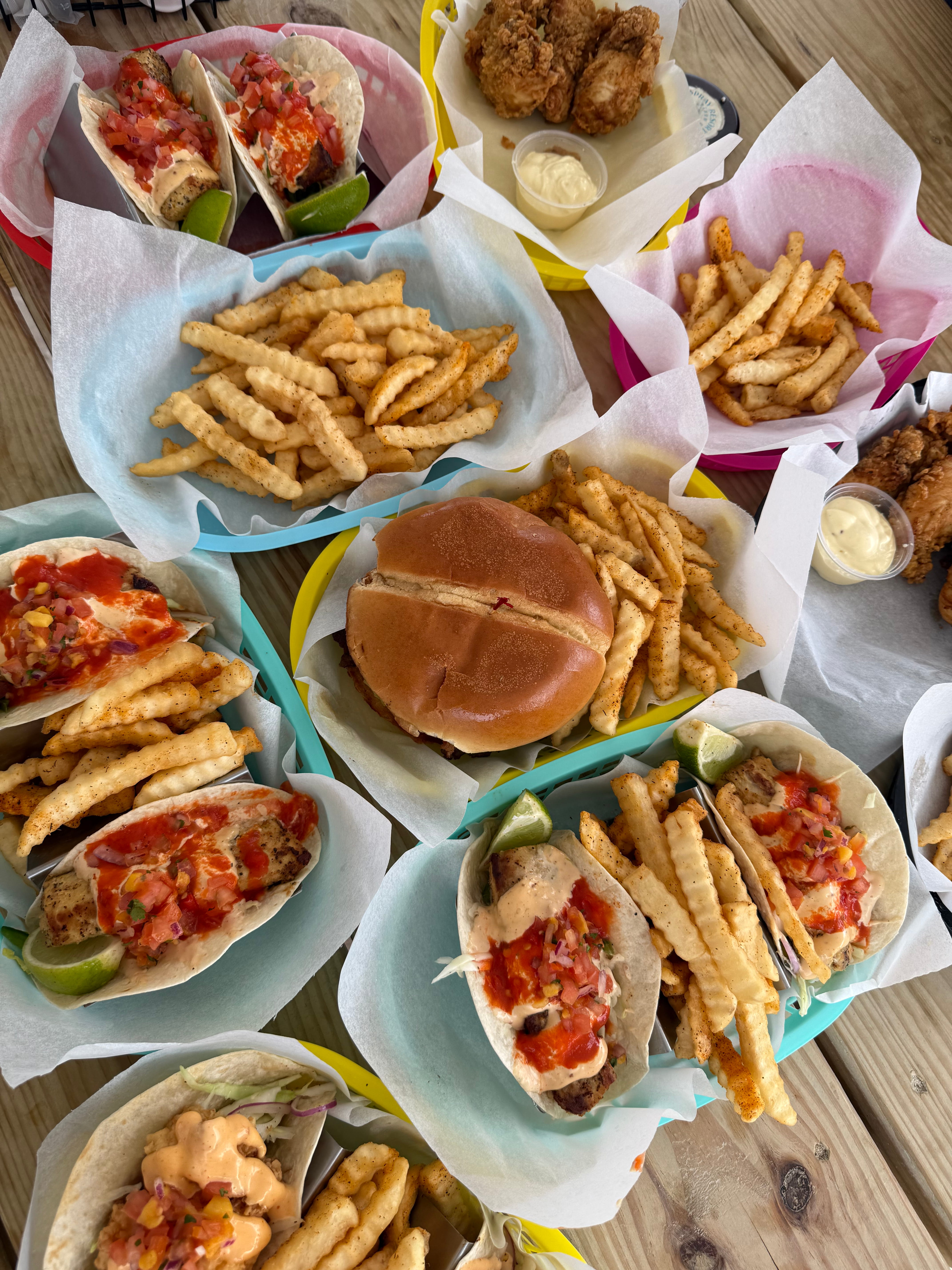 Table with baskets of grilled fish tacos topped with salsa, crinkle-cut fries, fried chicken, and a hamburger.