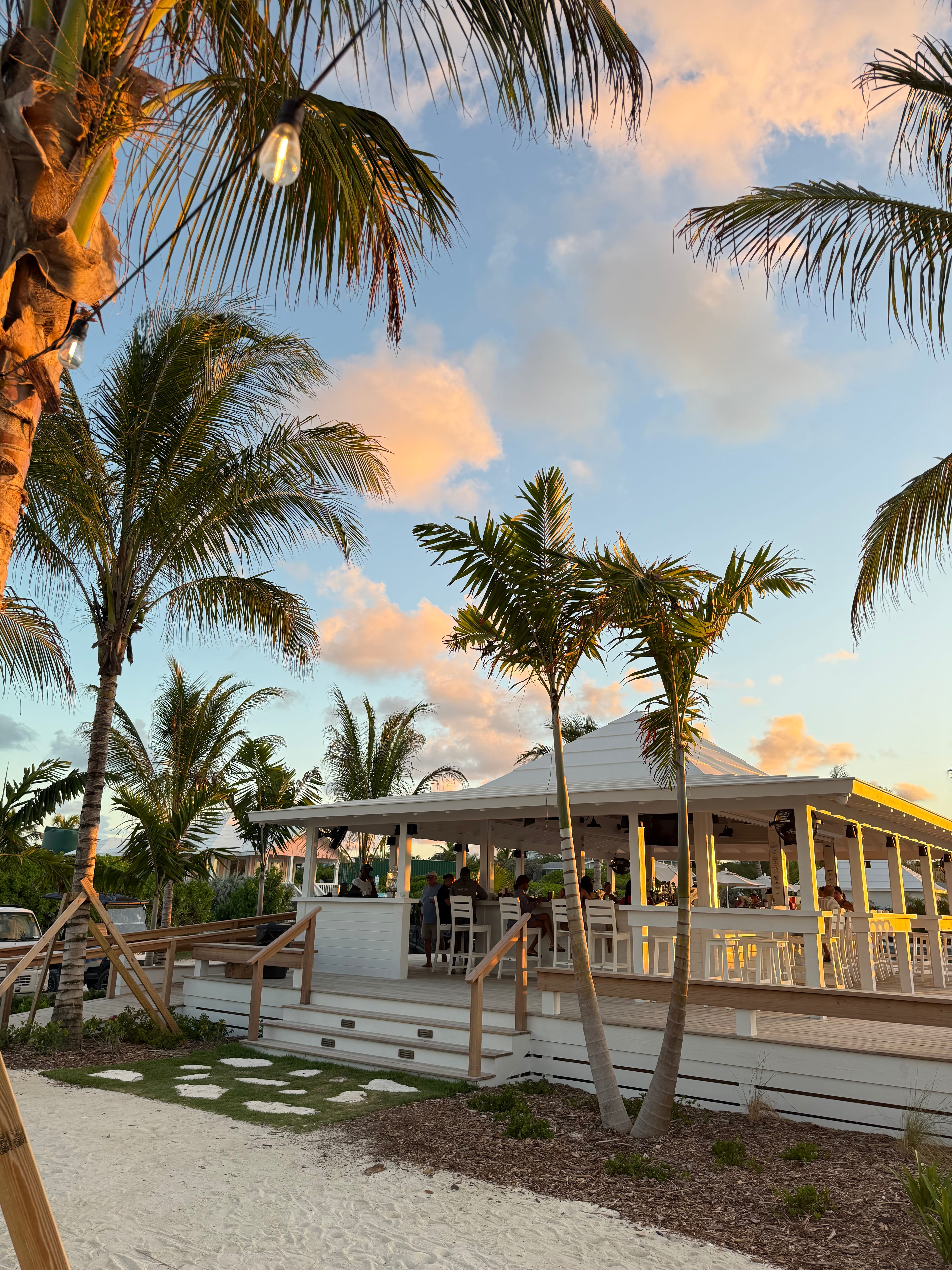 Open-air, white wooden bar and dining area surrounded by palm trees at sunset with people seated inside.