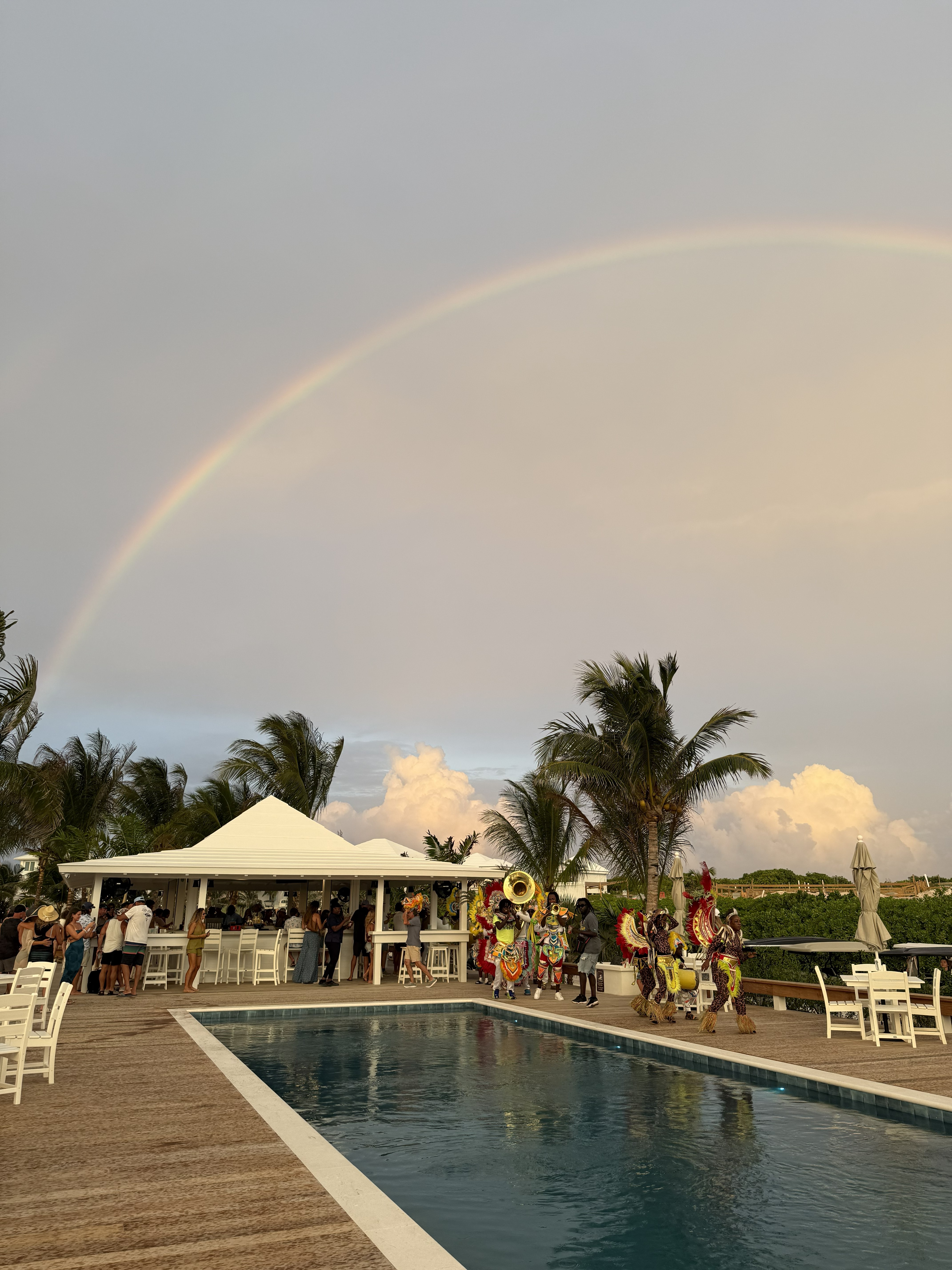 Evening poolside party with string lights, palm trees, and groups of people socializing near a lit swimming pool by the waterfront.