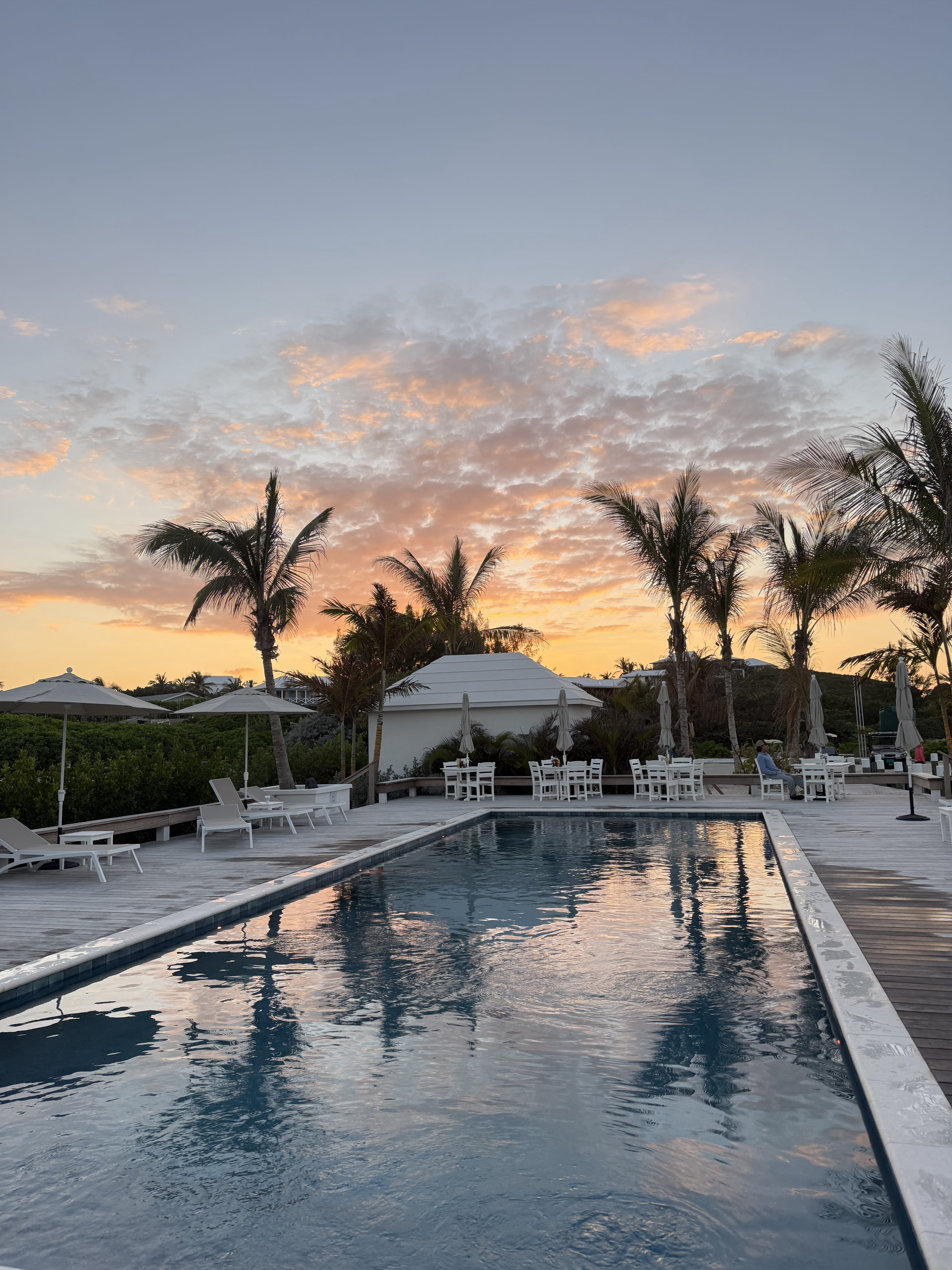Outdoor swimming pool at sunset with palm trees and lounge chairs around a wooden deck.
