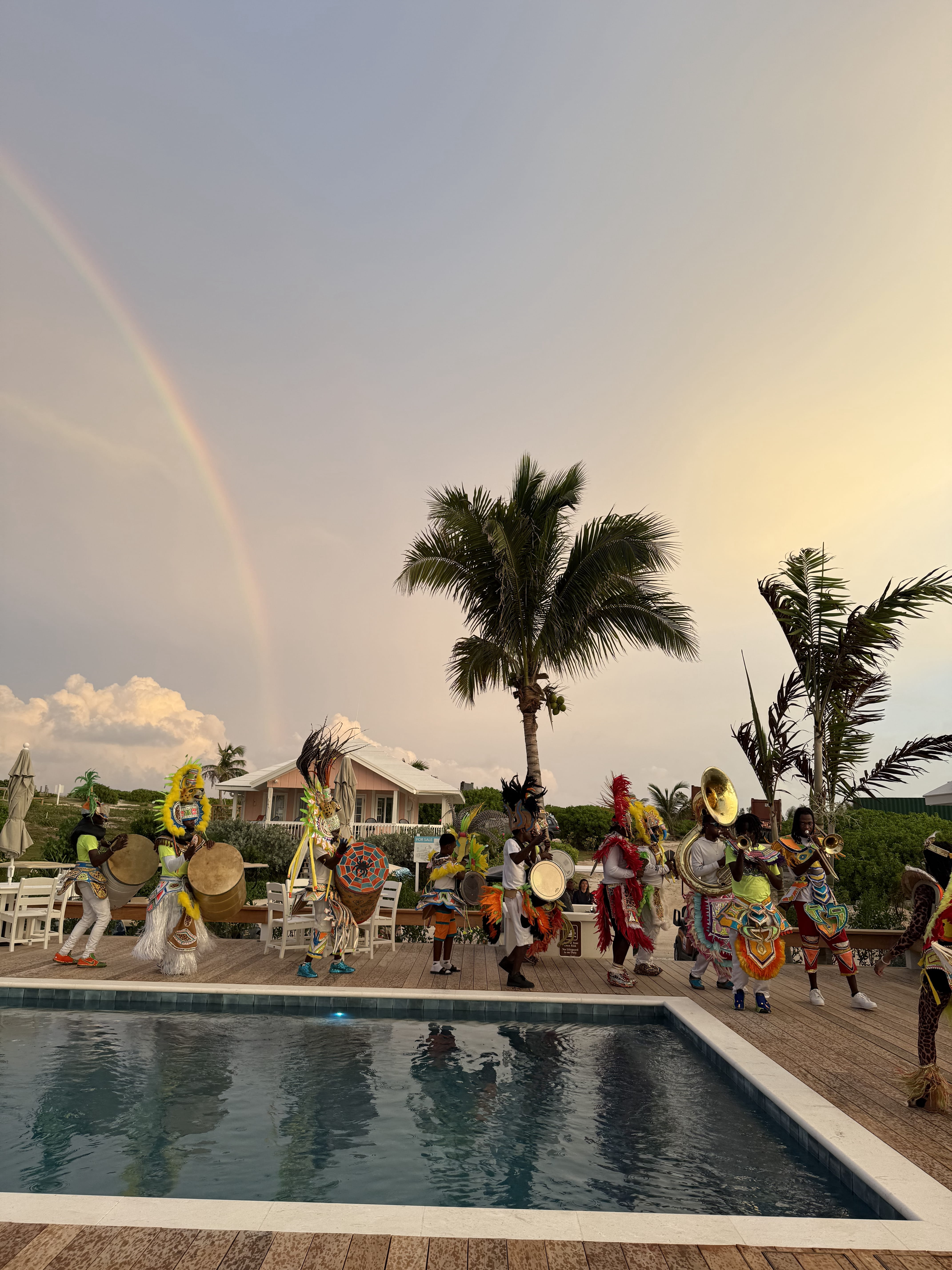 Group of musicians in vibrant traditional costumes playing instruments on a wooden deck beside a pool with a palm tree and a faint rainbow in the sky.