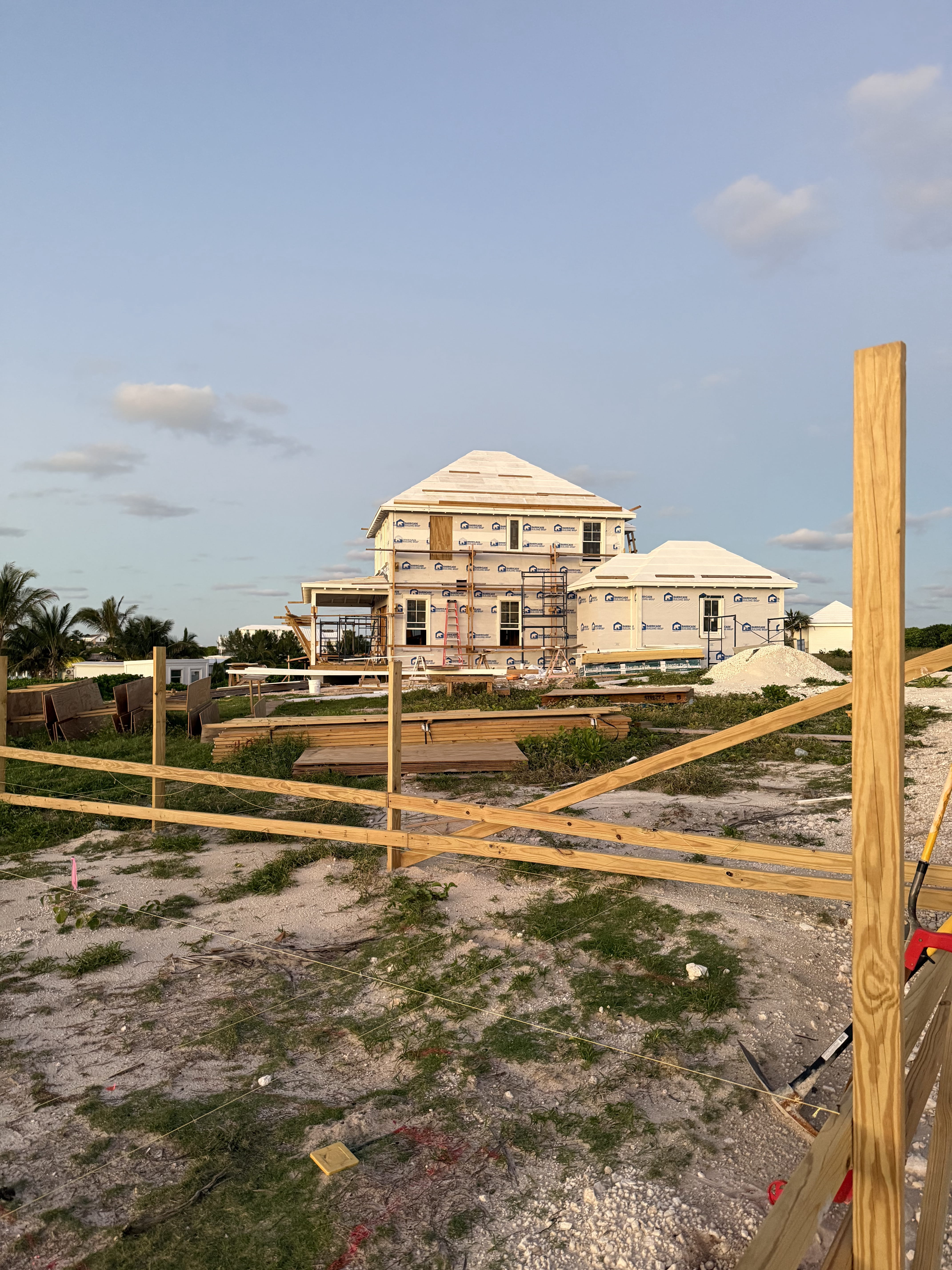 Partially built two-story house with white wrap on exterior, surrounded by wooden construction framework and building materials on sandy ground.