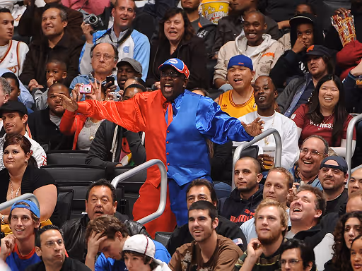 Energetic sports fan in a half red, half blue suit cheering among a diverse crowd in stadium seats.