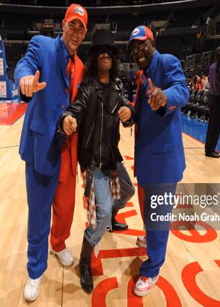 Three men posing on a basketball court, two in blue suits and caps pointing, and one in the middle wearing a black hat, sunglasses, leather jacket, and ripped jeans.