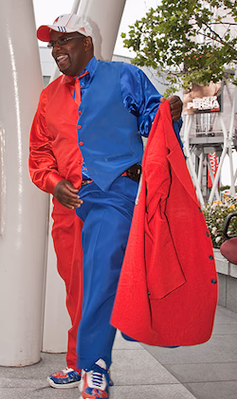 Smiling man wearing a half red and half blue outfit, holding a red jacket and wearing a white cap.