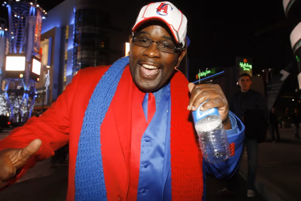 Man wearing a red suit, blue vest, and red and blue scarf smiling and holding a water bottle at night in an urban area.