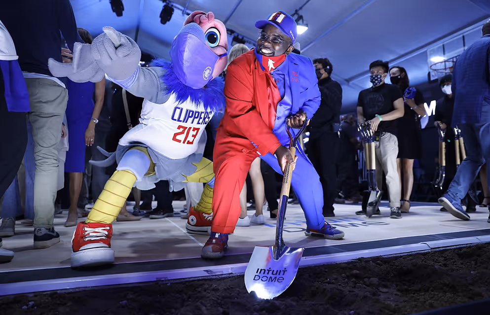 Man in a red and blue suit holding a shovel labeled Intuit Dome, next to a person in a Clippers mascot costume, surrounded by people indoors.