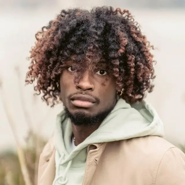 Young man with curly hair dyed with reddish tips wearing a beige jacket over a light green hoodie, looking directly at the camera.