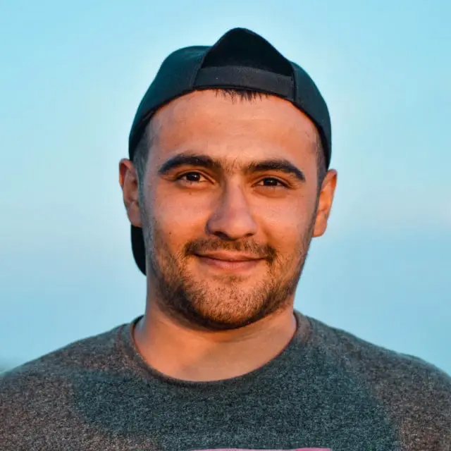 Smiling man wearing a backward black cap and a gray shirt against a clear blue sky.