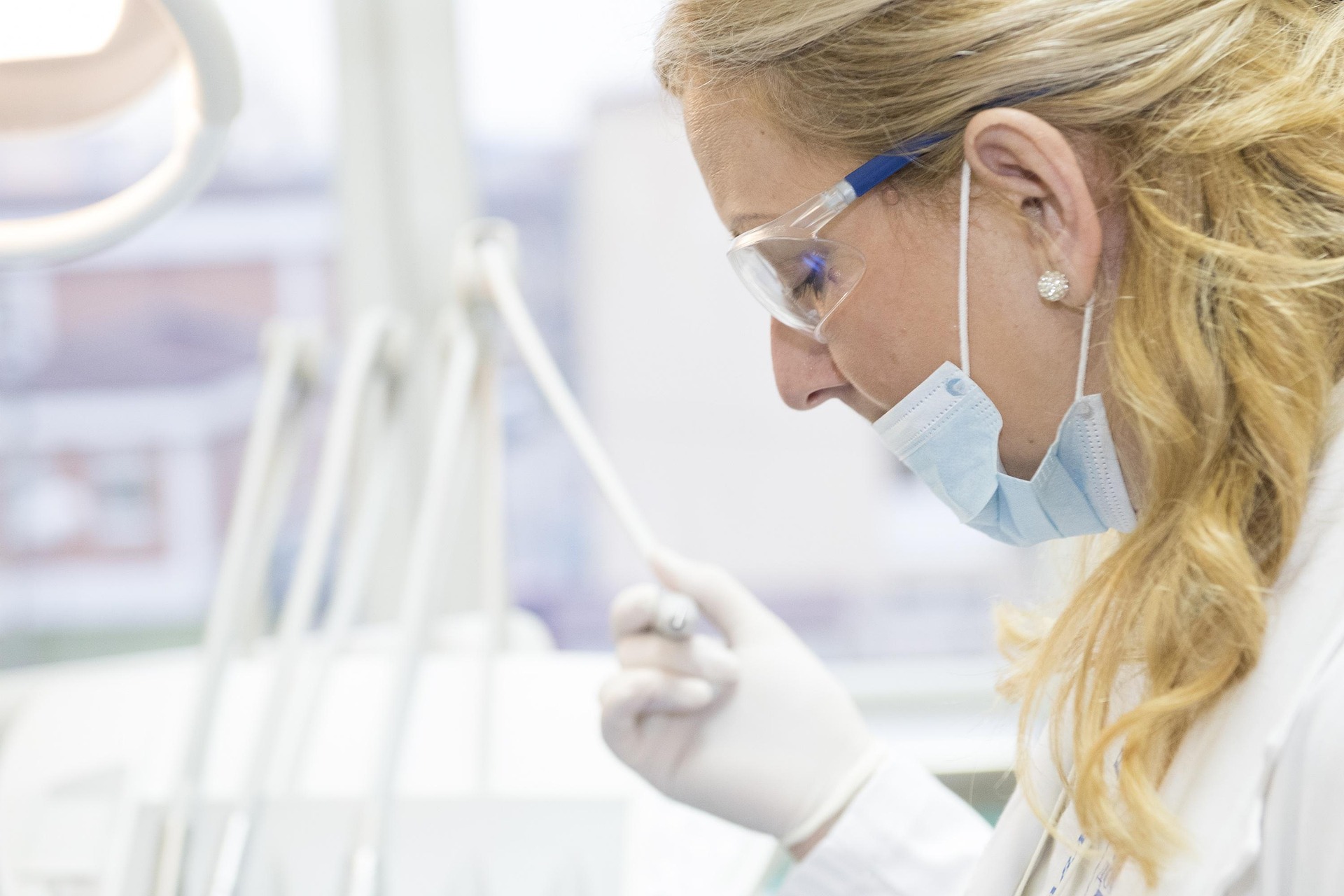Female medical professional wearing protective glasses, mask, gloves, and white coat working in a clinical setting.