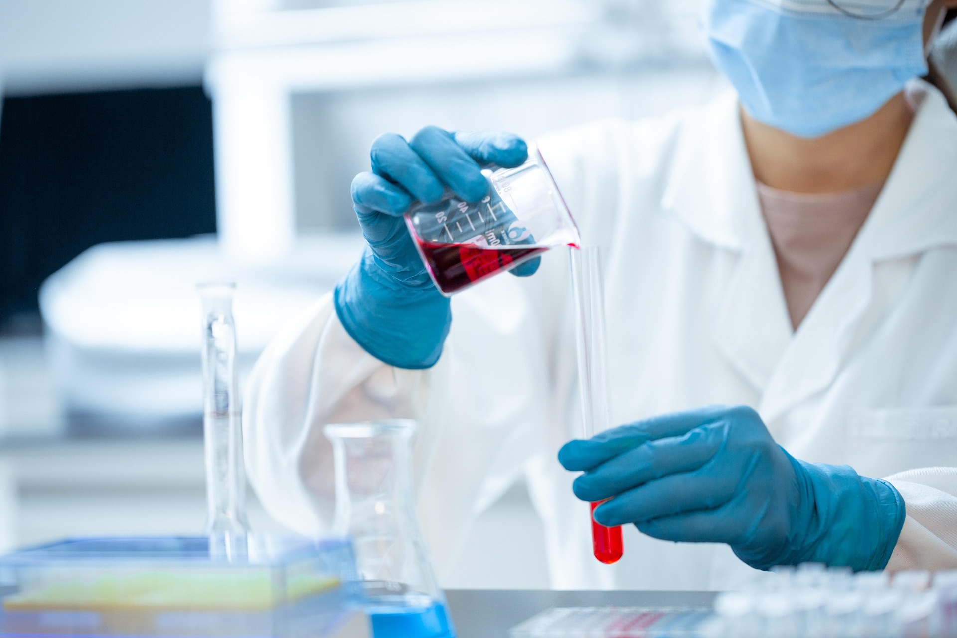Scientist in a lab coat and blue gloves pouring red liquid from a beaker into a test tube.