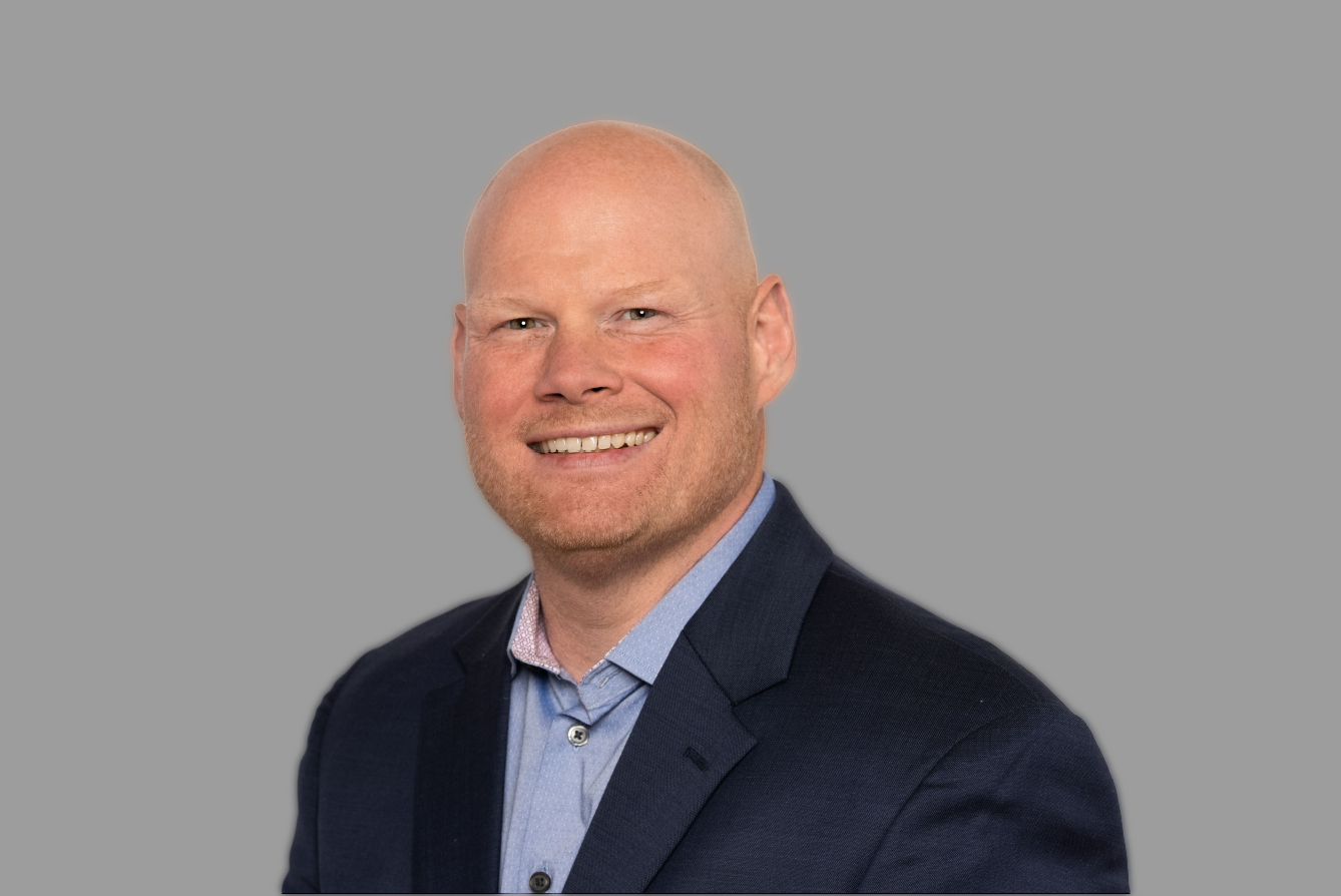 Smiling bald man wearing a blue shirt and dark blazer against a grey background.
