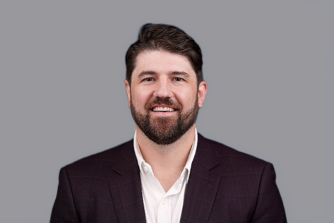 Smiling man with short brown hair and beard wearing a white shirt and dark blazer against a gray background.