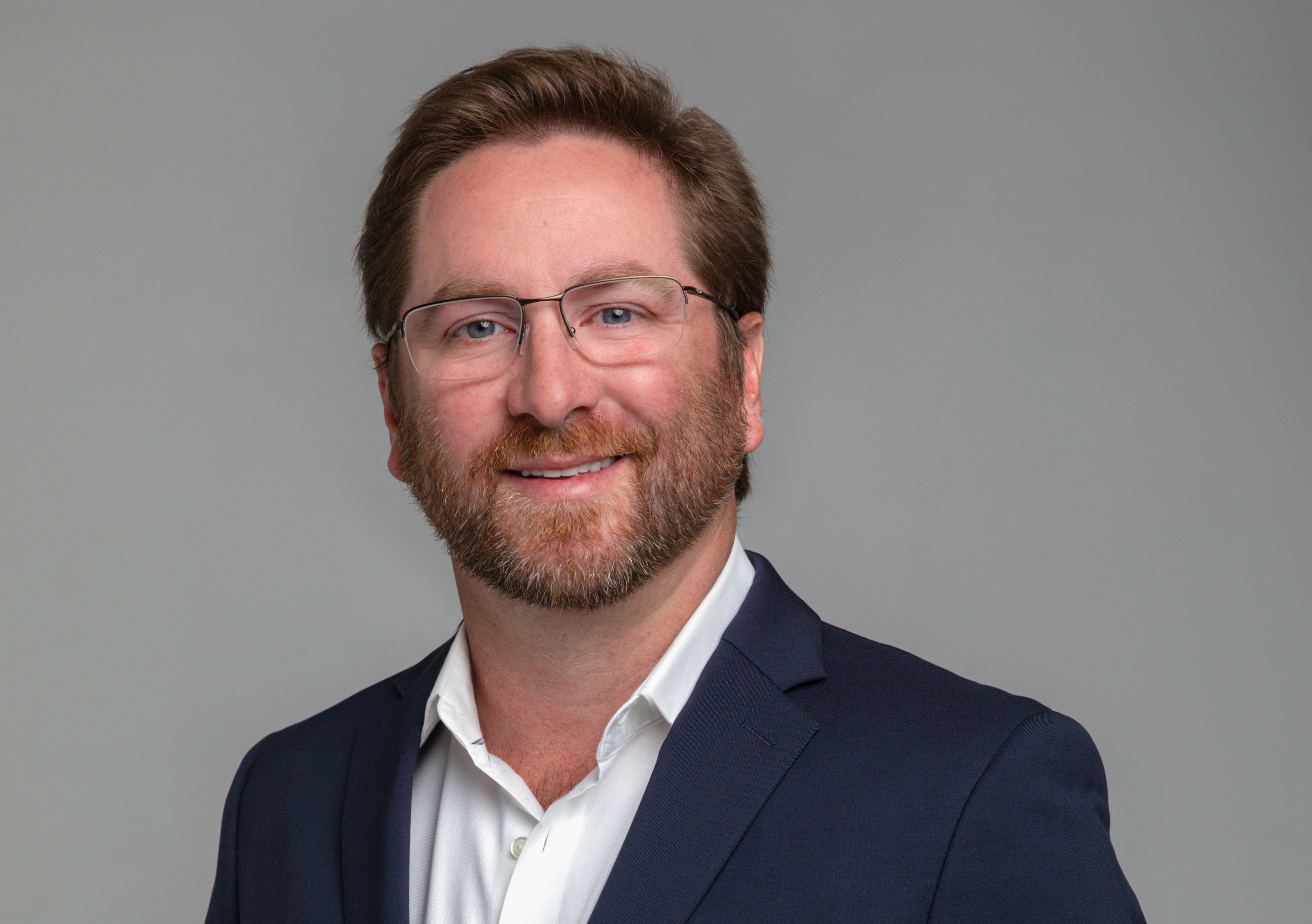 Smiling man with glasses, beard, and brown hair wearing a navy blazer and white shirt against a gray background.