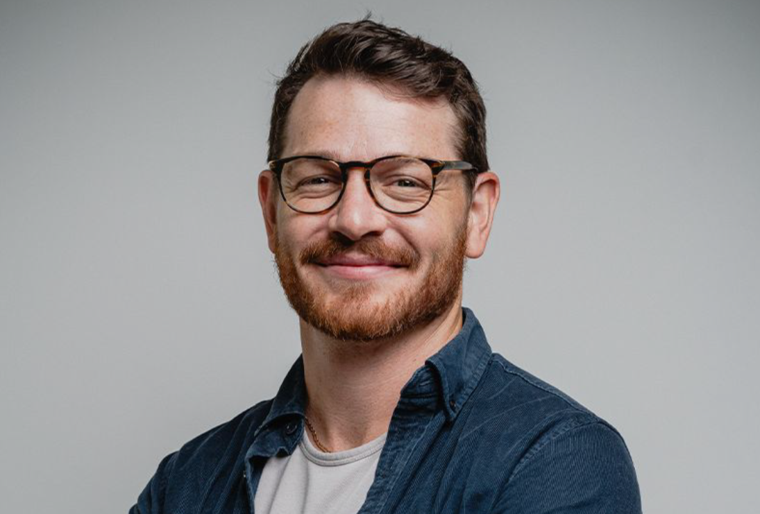 Smiling man with glasses and short brown hair wearing a blue shirt and gray t-shirt.