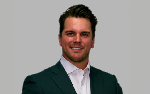 Smiling man with dark hair wearing a dark suit jacket and white dress shirt against a plain gray background.