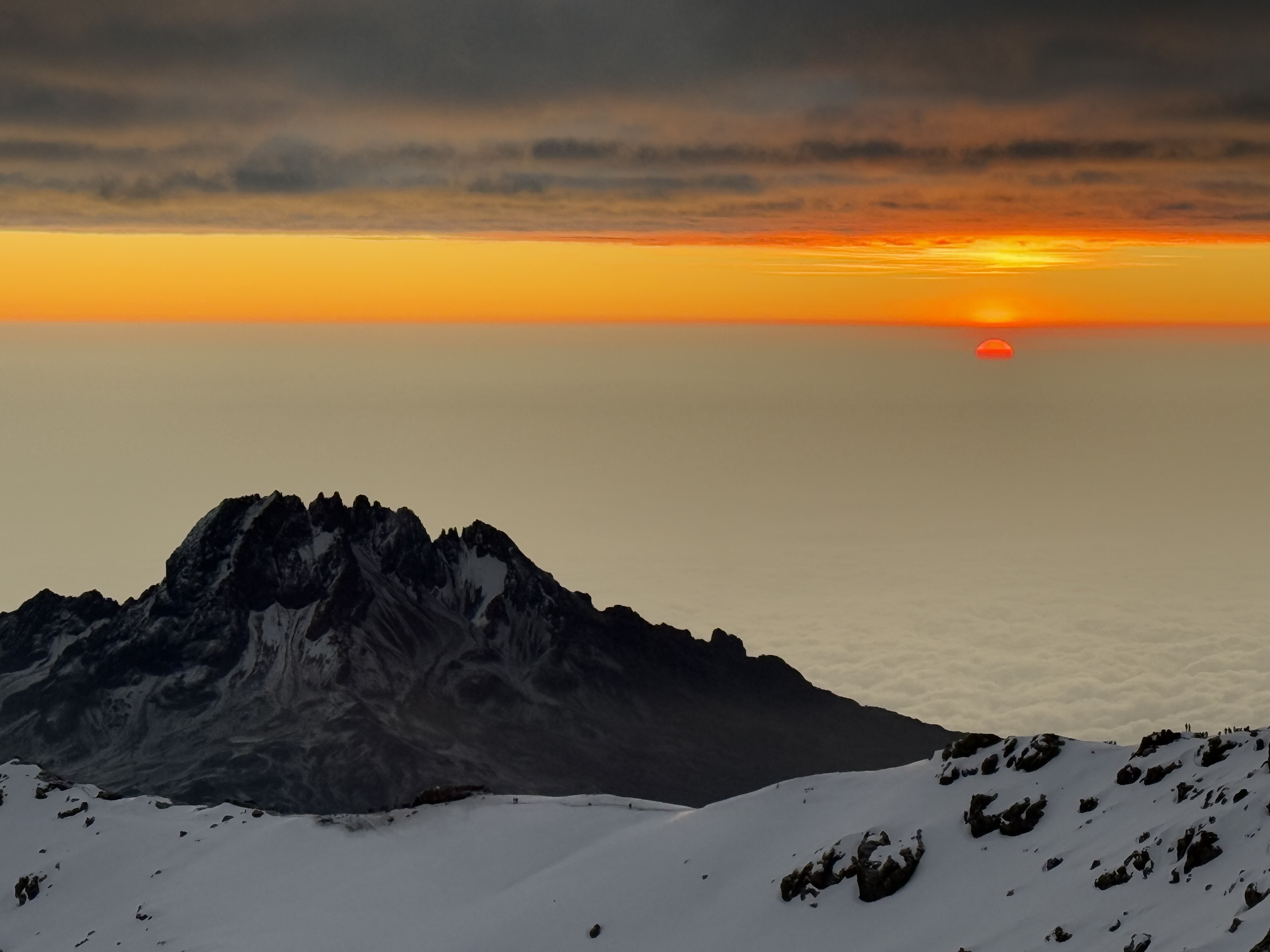 Snow-covered mountain ridge at sunrise with the sun partially visible above a layer of clouds.