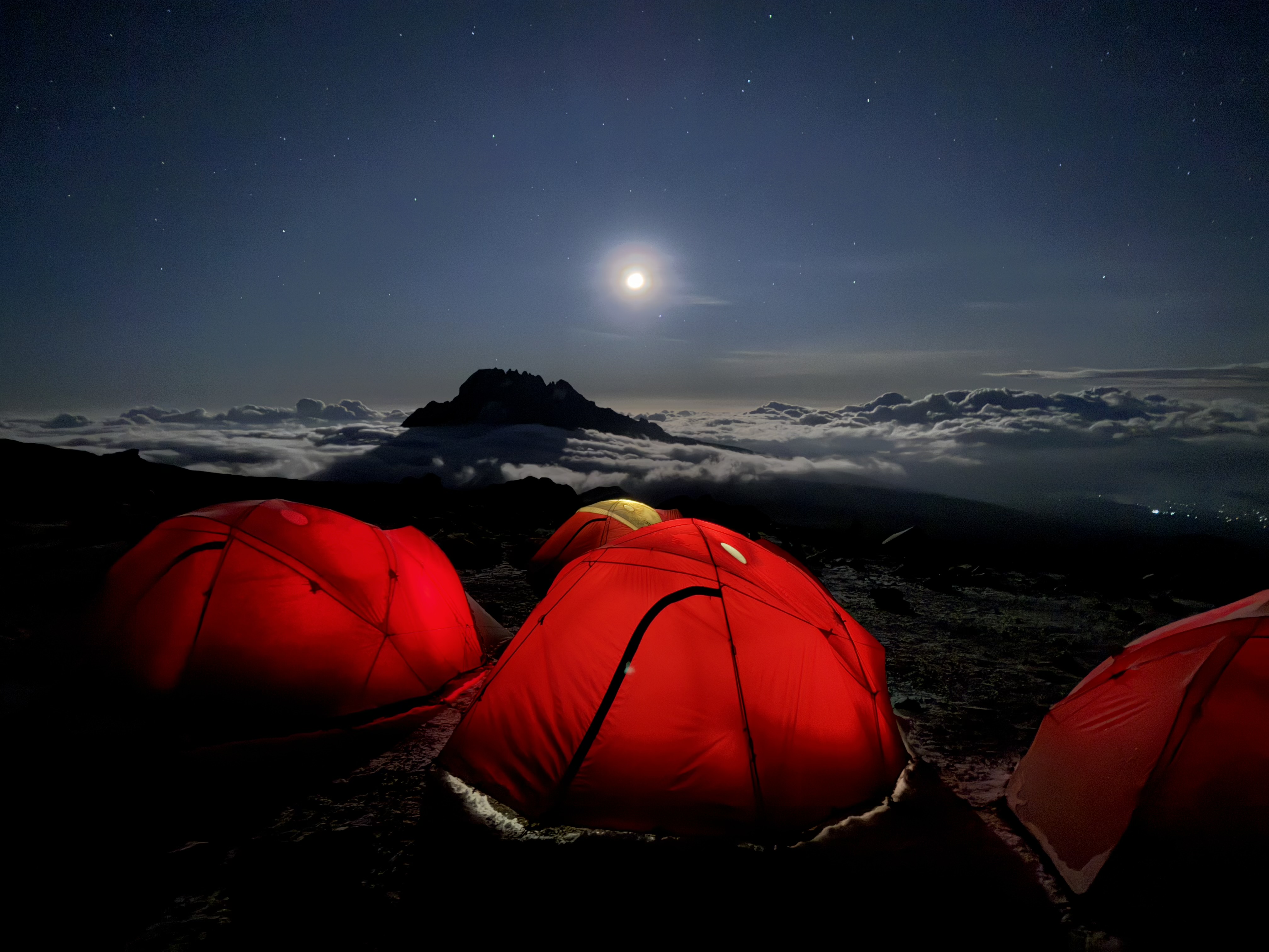 Red tents illuminated under a starry night sky with a full moon above a mountain surrounded by clouds.