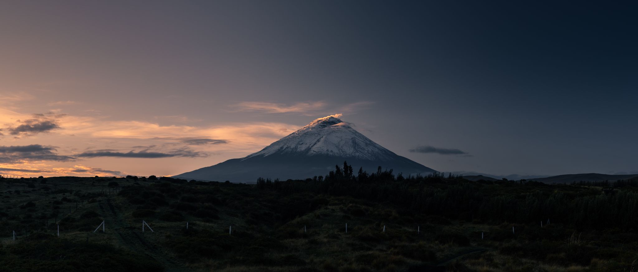 Snow-capped mountain under a darkening sky at sunset with grassy hills in the foreground.
