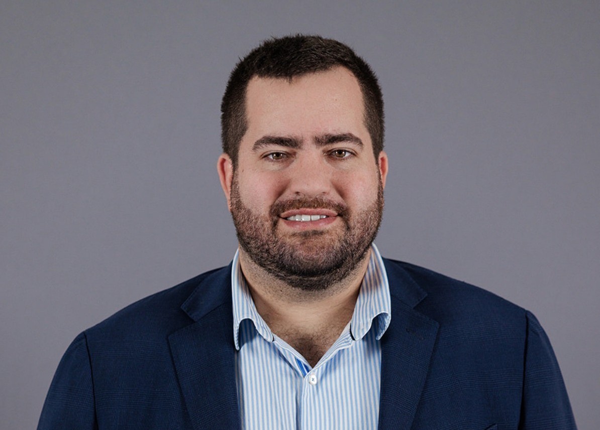 Bearded man wearing a blue blazer and light blue striped dress shirt against a plain gray background.