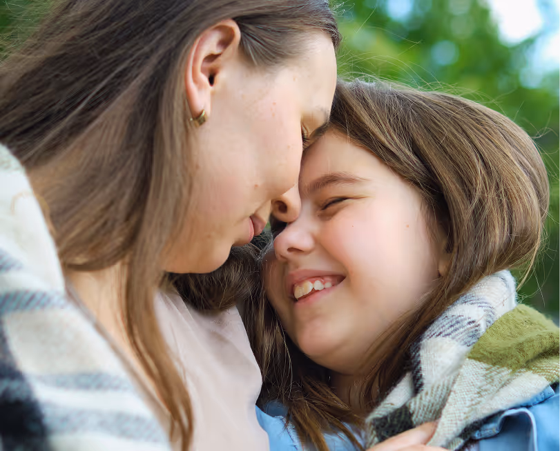 Close-up of a woman and a young girl smiling affectionately with their foreheads touching, wrapped in a plaid blanket outdoors.