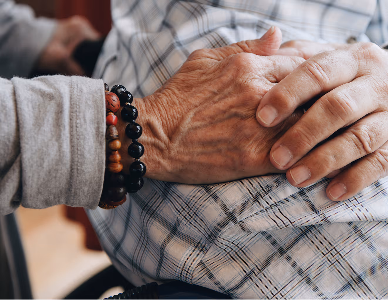 Close-up of one person holding the wrinkled hands of another, who is wearing a plaid shirt and wood bead bracelets.