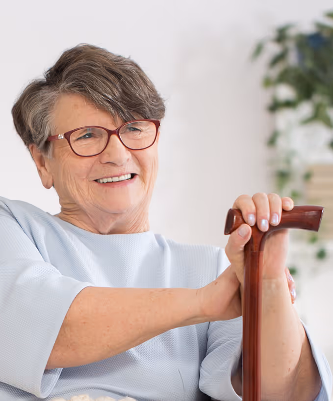 Smiling elderly woman with short gray hair and glasses holding a wooden cane indoors.